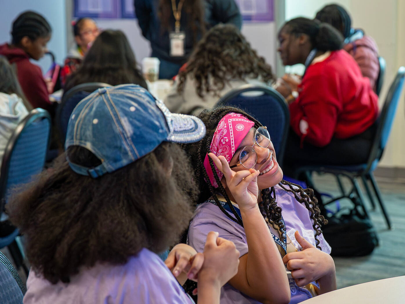 A young person wearing a pink bandana and glasses smiles while making a peace sign at a table, surrounded by others in a group setting.